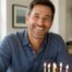 Smiling man celebrating his birthday at home, seated at a table with a birthday cake and wrapped gift in a bright, sunlit room.