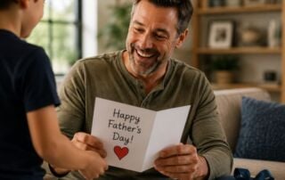 Father smiles while reading a handmade Father's Day card from his child, with a wrapped gift nearby in a cozy living room.