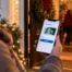 Person holding a smartphone displaying a $25 Starbucks digital gift card outside a festive holiday gathering.